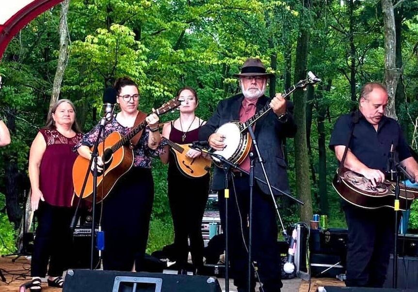 6 musicians on stage playing instruments with trees in the background